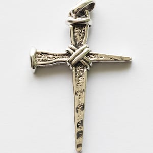 May include: A silver-colored cross pendant with a nail design. The cross features a textured surface and a loop at the top for a chain. The nail is wrapped with a textured band, creating a unique religious symbol.