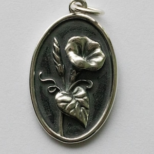 May include: A silver oval pendant featuring a detailed, three-dimensional design of a morning glory flower with a leaf and stem.