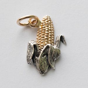 Könnte beinhalten: Ein goldfarbener Mais-Anhänger mit einer silberfarbenen Hülle. Der Mais ist mit detaillierten, strukturierten Körnern und einer Schlaufe zum Befestigen an einer Halskette oder einem Armband versehen. Der Anhänger ist eine Miniaturdarstellung eines Maiskolbens.