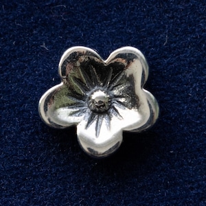 May include: A silver flower charm with five petals and a raised center. The charm is a simple and elegant design.