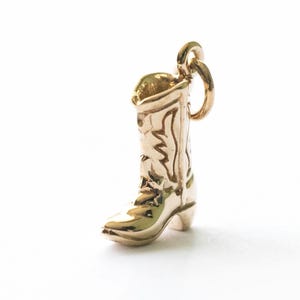 Könnte beinhalten: Ein goldfarbener Anhänger in Form eines Cowboy-Stiefels. Der Stiefel hat aufwendige Details, einschließlich eines dekorativen Musters auf dem Schaft und einem kleinen Absatz. Der Anhänger hat oben eine Schlaufe zum Befestigen an einer Halskette oder einem Armband.