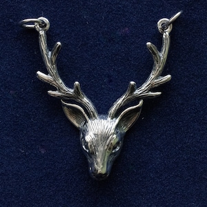 May include: A silver-coloured pendant depicting a deer's head with antlers. The pendant has a detailed, textured surface and two small loops at the top for a chain. The deer's head is centered, with the antlers extending upwards.