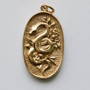 以下が含まれることがあります： 金色の楕円形ペンダント。蛇が花に巻き付いた詳細なレリーフが特徴です。背景はテクスチャ加工された斑点模様です。チェーンを通すための小さなループが上部にあります。