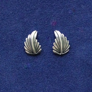 May include: A pair of silver-coloured leaf-shaped stud earrings. Each earring has a detailed, fan-like design with multiple curved lines, creating a textured appearance. The earrings are displayed on a dark blue surface.