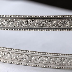 Könnte beinhalten: Zwei silberfarbene dekorative Metallstreifen mit einem verzierten Blumen- und Scroll-Design. Die Streifen haben einen Perlenrand und sind gebogen, was darauf hindeutet, dass sie Teil eines größeren dekorativen Elements sind. Das Design ist detailliert und elegant.