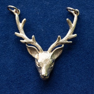 May include: A gold-coloured pendant shaped like a stag's head, featuring detailed antlers. The pendant has two small loops at the top for a chain. The stag's head is set against a dark blue backdrop.