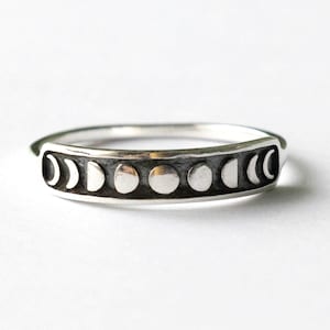 Peut inclure: Une bague en argent représentant les phases de la lune. La bague a un bandeau foncé et texturé, affichant la progression de la lune du croissant à la pleine lune. La bague est posée sur un fond blanc.