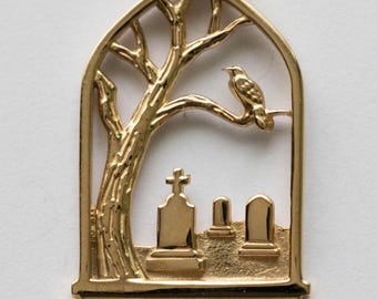 May include: A gold-colored pendant featuring a tree with a perched bird, set within an arched frame. The scene includes a graveyard with three tombstones. The pendant has a loop for a chain.