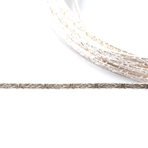 Könnte beinhalten: Ein silberfarbenes Armband mit einem sich wiederholenden floralen Muster. Das Armband wird vor einem weißen Hintergrund gezeigt. Das Design besteht aus kleinen, symmetrischen floralen Mustern, die miteinander verbunden sind.