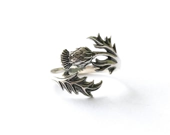 May include: A silver-toned ring featuring a detailed thistle design. The ring wraps around the finger with the thistle bloom at the center and stylized leaves on either side. The ring is set against a white background.