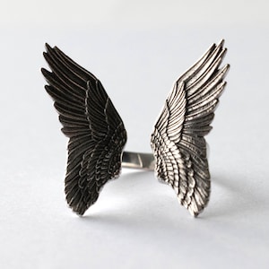 May include: A silver-toned ring featuring a detailed design of two open wings. The wings are intricately crafted with individual feather details, creating a textured appearance. The ring's design is symmetrical, with the wings extending outwards from the band.