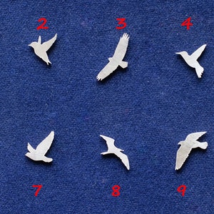 Op de afbeelding: Tien witte metalen vogeluitsparingen op een blauwe stoffen achtergrond. De vogels zijn in verschillende poses en maten. De uitsparingen zijn genummerd van 1 tot en met 10.
