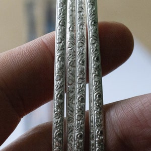 May include: Four silver bangles with intricate floral designs. The bangles are stacked on top of each other and have a shiny finish.