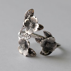 May include: A silver ring with a floral design. The ring features three open flowers and leaves, wrapping around the finger. The flowers and leaves extend outwards. The ring is made of a shiny metal, with a detailed floral pattern.