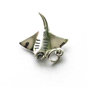 Op de afbeelding: Een zilveren manta rog bedel met een gebogen staart en vleugelachtige vinnen. De bedel heeft een kleine lus om aan een ketting of armband te bevestigen. De tekst "925 n" is zichtbaar.