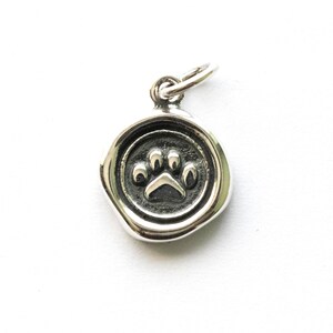 May include: A small, circular silver charm featuring a paw print. The charm has a raised paw print design in the centre, encircled by a border. It has a loop at the top for attaching to a necklace or bracelet.