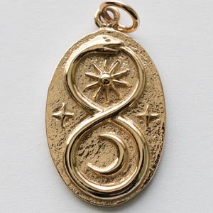 May include: Gold-coloured pendant with an oval shape. The pendant features an ouroboros design, a sunburst, a crescent moon, and two stars. The pendant has a textured surface and a loop at the top for a chain.