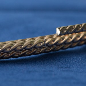 May include: A close-up of a gold-colored braided metal band. The band is twisted and has a textured surface.