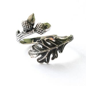 May include: A silver ring with an oak leaf and acorn design. The ring wraps around the finger, with an oak leaf on one end and two acorns on a branch on the other. The ring is set against a white background.