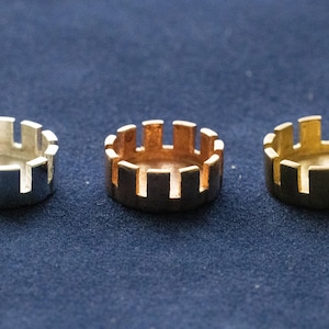 Op de afbeelding: Drie metalen ringen, een zilveren, een koperen en een gouden, met een decoratief, opengewerkt ontwerp. De ringen zijn in een rij op een blauw oppervlak gerangschikt.