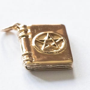 May include: A gold-toned metal charm shaped like a small book with a raised pentagram symbol on the front cover. The charm features a detailed spine with horizontal bands and a small jump ring attached to the top for hanging as a pendant.