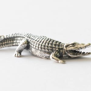 Könnte beinhalten: Eine detaillierte silberne Alligatorfigur, die auf einer weißen Oberfläche liegt. Der Alligator hat ein offenes Maul, das Zähne zeigt, und einen strukturierten Körper. Die Figur ist eine detaillierte Darstellung des Reptils.
