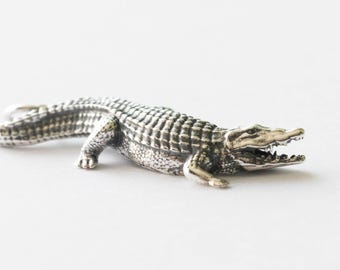 May include: A detailed silver alligator figurine, lying on a white surface. The alligator has an open mouth, revealing teeth, and a textured body. The figurine is a detailed representation of the reptile.