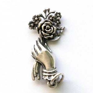Peut inclure: Pendentif argenté représentant une main tenant un bouquet de fleurs. Le bouquet comprend une rose et de petites fleurs. La main est ornée d'un bracelet de cheville détaillé et d'une boucle en bas. Le pendentif est probablement en argent sterling.