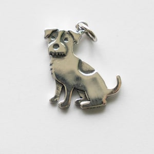 May include: A silver charm in the shape of a dog. The dog is sitting with its head turned to the side and its tail curled up.