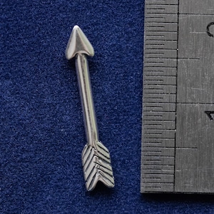 Könnte beinhalten: Ein silberner Pfeil-Anhänger mit einer spitzen Spitze und einem detaillierten Federdesign. Der Anhänger ist etwa 2,5 cm lang und wird neben einem Lineal gezeigt.