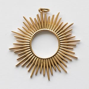 May include: A gold-colored pendant in the shape of a sunburst. The pendant features a central circular design with radiating spikes. A small loop is at the top for hanging. The pendant is made of a shiny, metallic material.