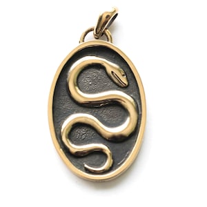 May include: Gold-coloured pendant with an oval shape. The pendant features a raised, stylised snake design in gold against a dark background. The snake is coiled within the oval frame, and the pendant has a loop for a chain.