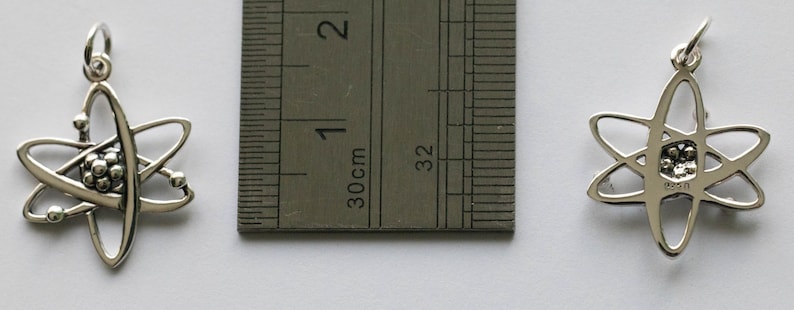 K&ouml;nnte beinhalten: Zwei silberfarbene Anh&auml;nger in Form eines Atoms. Die Anh&auml;nger haben einen Durchmesser von etwa 2,5 cm. Einer der Anh&auml;nger ist nach oben, der andere nach unten gerichtet. Ein Lineal ist zwischen den Anh&auml;ngern platziert und zeigt die Ma&szlig;e in Zentimetern an.