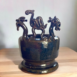 May include: A ceramic planter with a dark green glaze features three sculpted animal figures: a camel, a dragon, and a bird. The planter has a wide base and a rounded body.