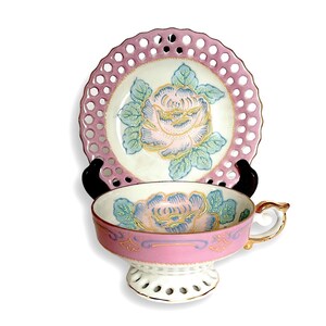 May include: A pink and white teacup and saucer set with a floral design. The saucer has a delicate openwork pattern around the edge. The teacup has a matching floral design and a delicate openwork base.
