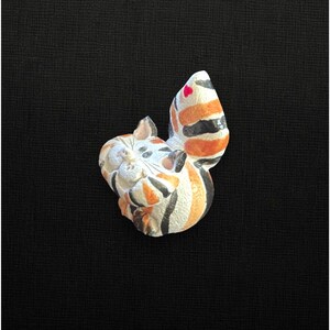 May include: A small, hand-painted ceramic cat figurine featuring a calico pattern with white, orange, and black stripes. The cat is depicted in a curled position with its tail raised, showcasing a tiny red heart detail on the tip of the tail.
