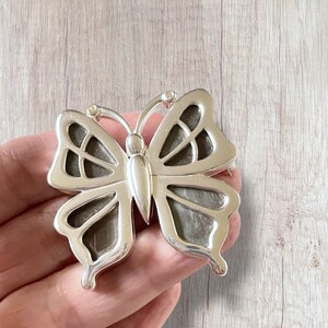 May include: Silver butterfly brooch with a detailed, intricate design. The wings are translucent and have a dark gray color.