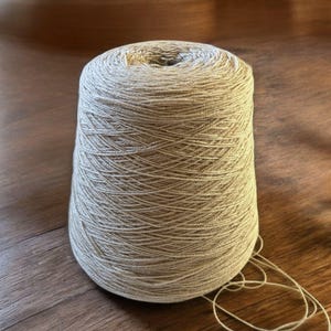 May include: A cone of beige yarn, wound tightly, with a loose end of yarn hanging off the side.