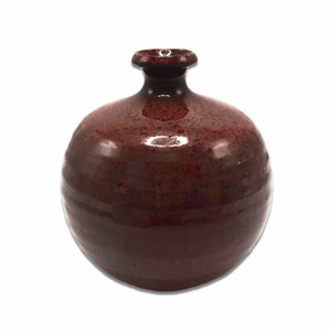 May include: A round, red ceramic vase with a textured glaze. The vase has a small opening at the top.