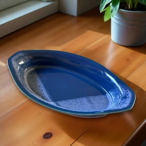 May include: A blue oval serving dish with a speckled interior and a glossy finish. The dish is set on a wooden surface, with a plant in a white pot visible in the background. The dish is approximately 12 inches long.