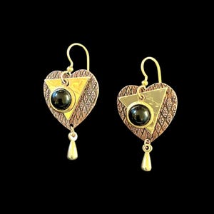 May include: A pair of heart-shaped earrings with a gold-colored hook. The earrings feature a triangle and a round black stone in the center. The earrings have a copper-colored border with a decorative pattern and a teardrop-shaped dangle.