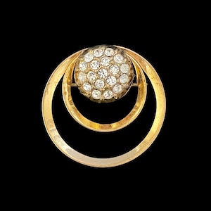 May include: A gold-toned brooch with a circular design. The center of the brooch is a cluster of clear rhinestones set in a smaller circle. The smaller circle is surrounded by a larger circle, creating a double circle design.