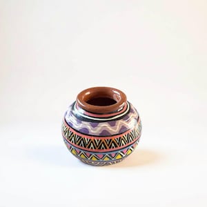 May include: A small, round ceramic pot with a wide opening and a brown rim. The pot is decorated with colorful geometric patterns in black, pink, yellow, and purple. The background is plain white.