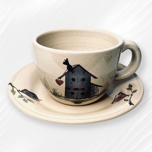 May include: A beige ceramic teacup and saucer set with a hand-painted design of a birdhouse with a black bird perched on top. The birdhouse has a red heart and blue accents.