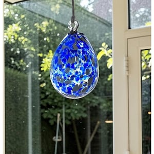 May include: A blue teardrop-shaped glass ornament with dark blue and black speckles, suspended by a black cord. The ornament is hanging in front of a window with a blurred green background.