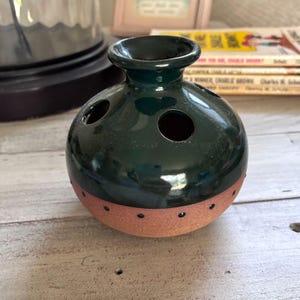 May include: A dark green ceramic vase with a round body and a narrow neck. The vase has three circular holes and a band of reddish-brown with small black dots around the base. The vase is on a light-colored wooden surface.