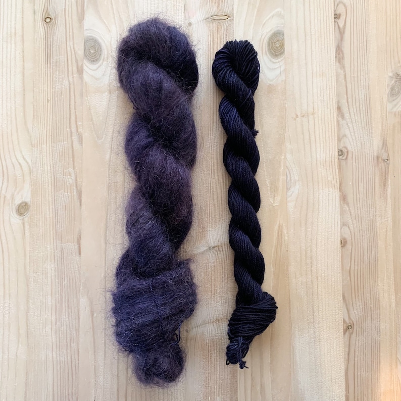 May include: Two skeins of yarn, one dark purple and one black, are displayed on a wooden surface.