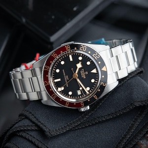 Peut inclure: Une montre-bracelet en acier inoxydable avec un cadran noir et une lunette rouge et noire. La montre a un bracelet argenté et le texte "TUDOR GENEVE GMT MASTER CHRONOMETER" est visible sur le cadran. La montre repose sur un tissu noir.