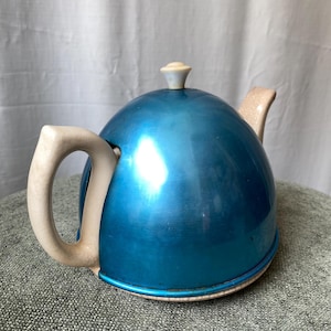 Puede incluir: Una tetera azul vintage con cuerpo redondeado y asa y pico crema. La tapa tiene un pequeño botón crema. La tetera, probablemente de cerámica o porcelana, descansa sobre una base clara.