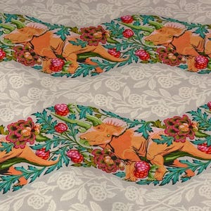 May include: White fabric with a repeating pattern of orange triceratops dinosaurs in a jungle setting with pink flowers and green leaves. The fabric has the text "Free Spirit Presents ROAR!" at the bottom.
